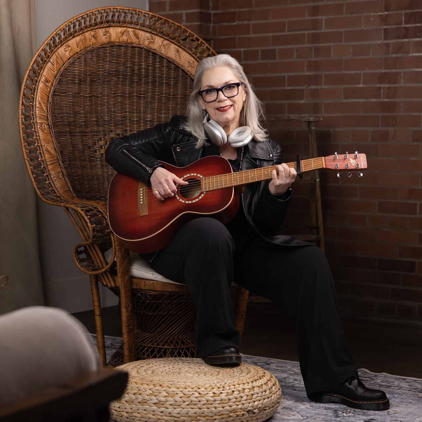 Woman wearing all black sits in a larger wicker chair holding her guitar