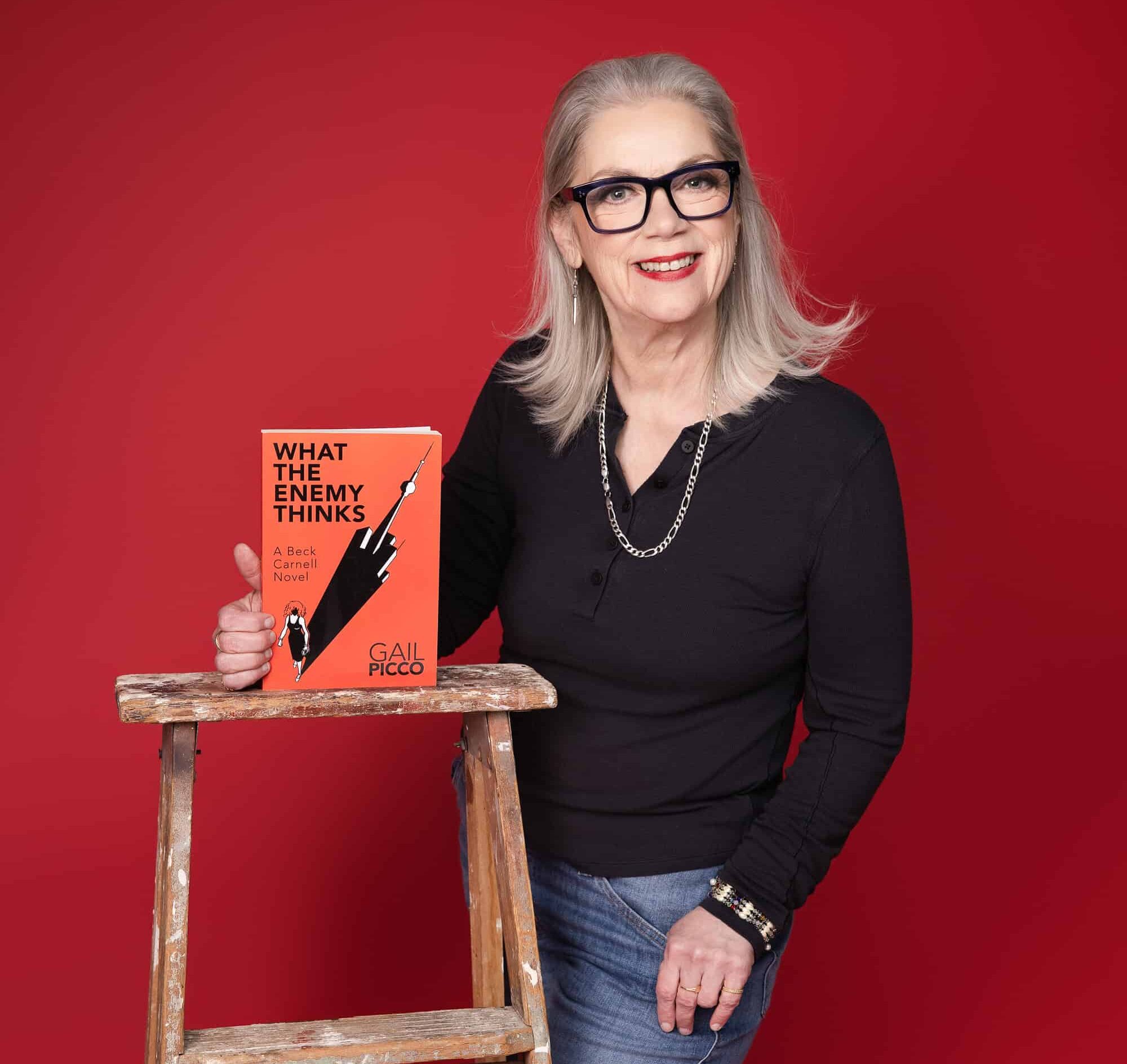 Woman in glasses and black sweater stands against a red background holding an orange book titled What the Enemy Thinks